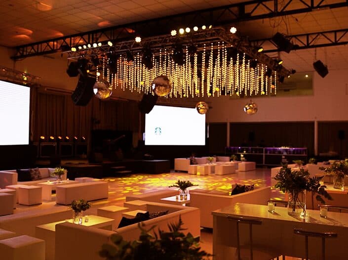 El salón montado en modo lounge y vacío, con sillones blancos y mesas bajas alrededor de la pista iluminada de amarillo, una estructura de truss con cortina de luces colgando del techo, bolas de espejo y dos pantallas grandes encendidas al fondo.