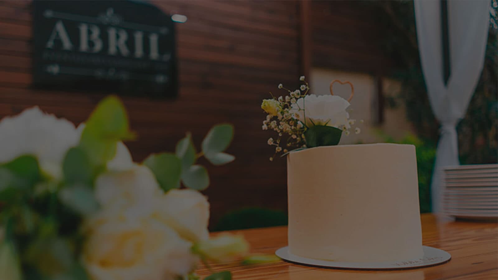 La torta de casamiento, lisa y blanca, sobre una mesa de madera, con flores en primer plano y el cartel del salón colgado en la pared de madera atrás.