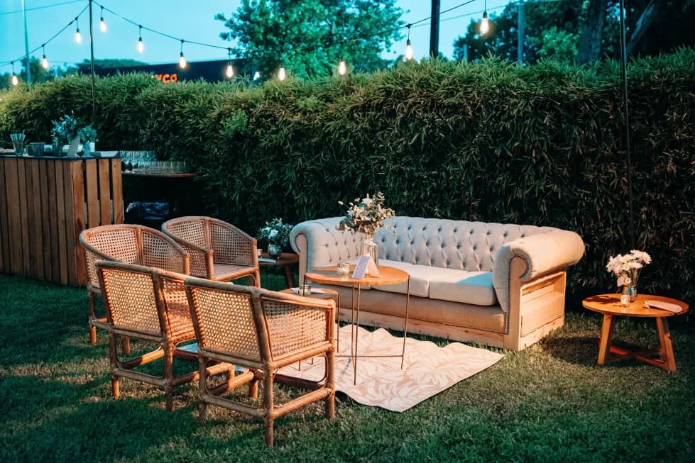 Un living armado sobre el césped al atardecer: sofá capitoné, sillones de mimbre y una mesita con flores, con la barra y una guirnalda de luces atrás.