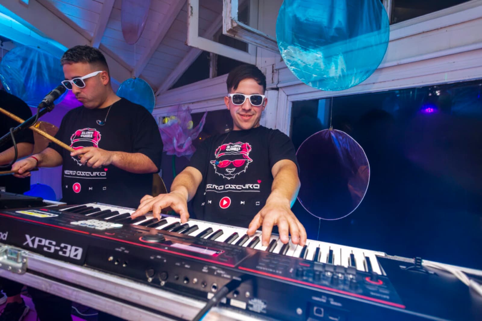 Dos integrantes de la banda tocando, uno en el teclado y otro en la percusión, con la fiesta iluminada de azul detrás.