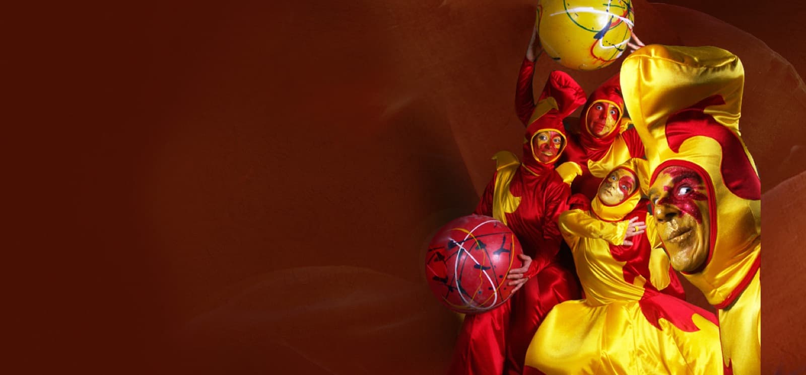 Un grupo de artistas vestidos de arlequín en rojo y amarillo, con máscaras y globos, sobre un fondo rojo.