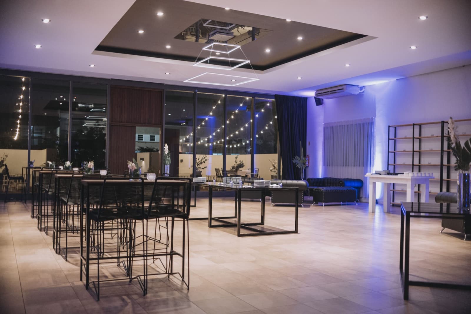 El sector de recepción vacío y encendido, con mesas altas de tapa negra y banquetas de hierro, una araña cuadrada de luz led en el cielorraso, sillones capitoné azules contra la pared y ventanales que dan a un patio con guirnaldas de luces.