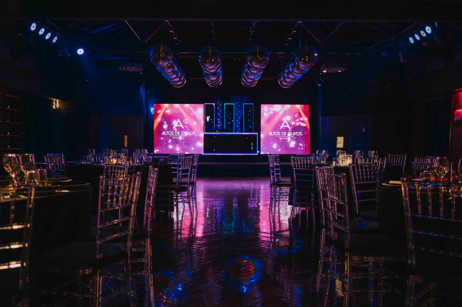 El salón principal armado para la fiesta, con mesas de mantel negro y sillas transparentes a los costados, bolas de espejos sobre la pista que refleja las luces y dos pantallas led con el logo de Altos de Olivos detrás de la cabina del DJ.