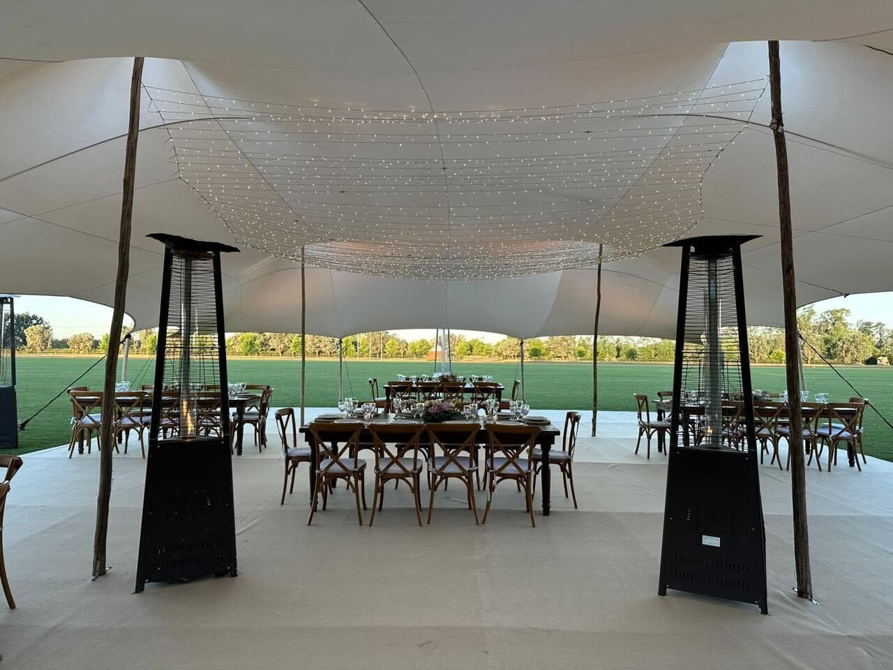 La carpa blanca montada sobre el campo, con una mesa larga de madera puesta en el centro, cortina de luces en el techo y el sembrado verde al fondo.