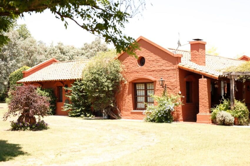 La casa de ladrillo a la vista con techo de tejas y galería, rodeada de plantas y con el parque adelante.