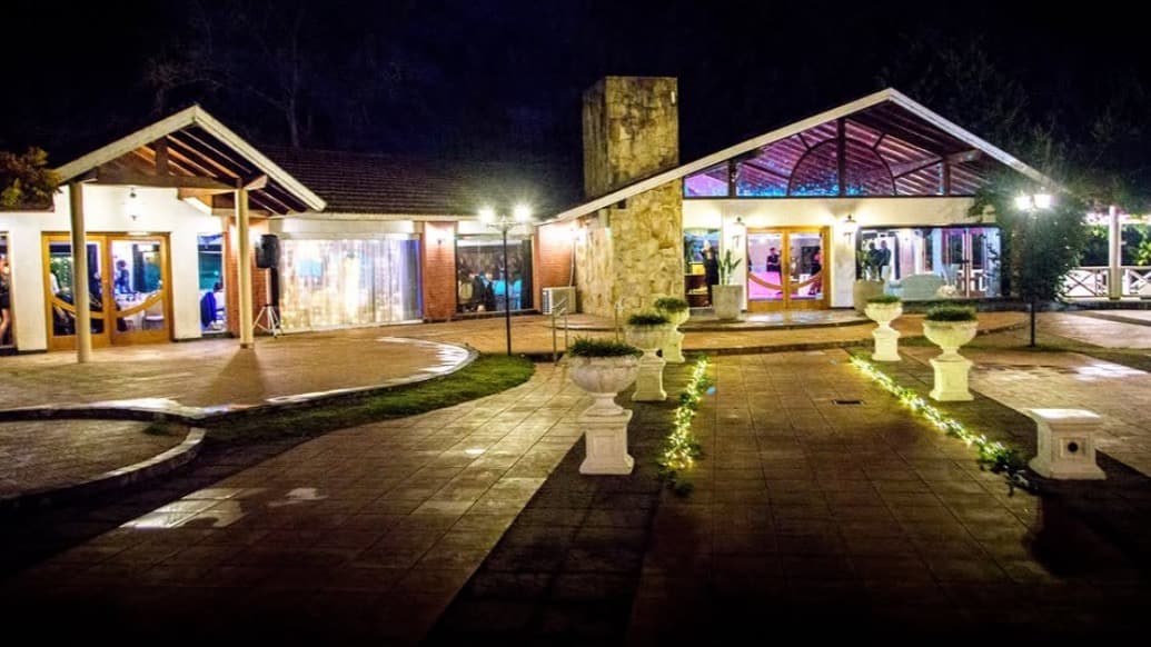 El complejo de noche desde el patio, con los dos salones de techo a dos aguas iluminados, macetones blancos sobre el piso de baldosas y guirnaldas de luces en los canteros.