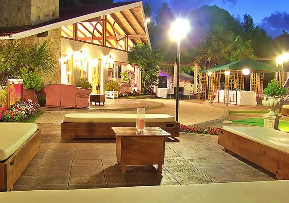 La galería del salón iluminada al anochecer, con los bancos de madera y los canteros del parque adelante.