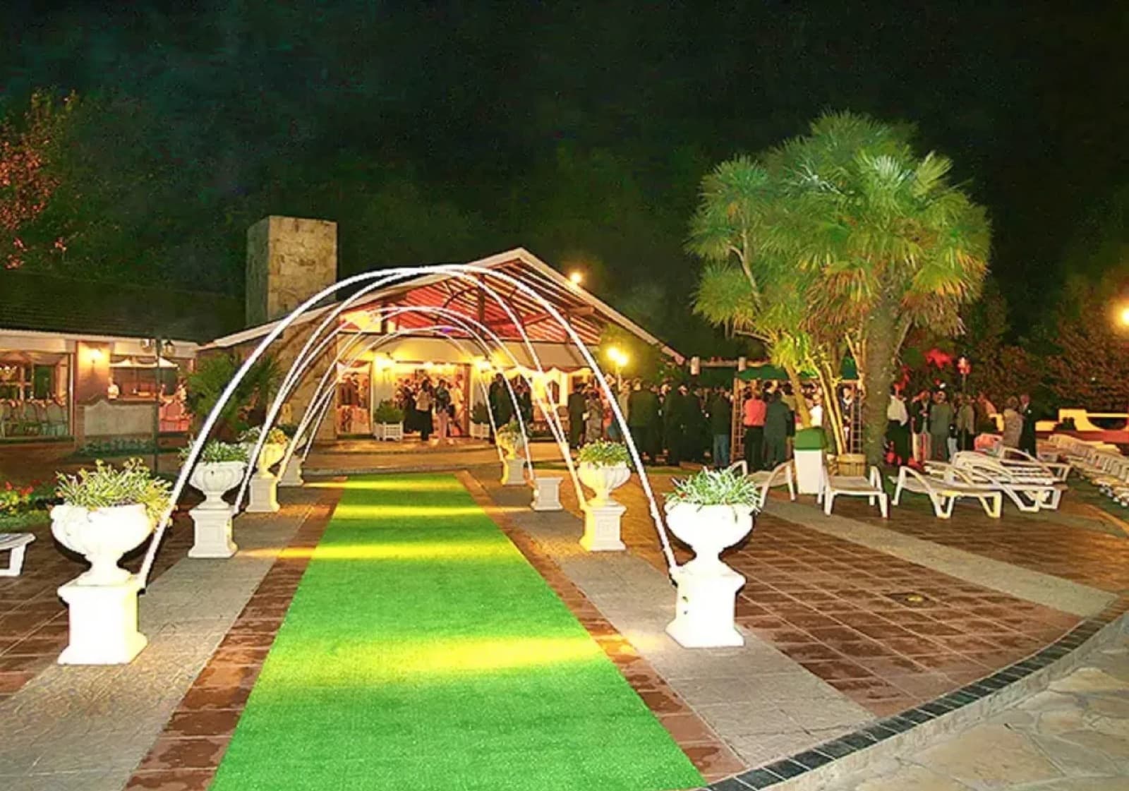 El camino de acceso al salón de noche, con arcos blancos sobre la alfombra verde y macetones a los costados.