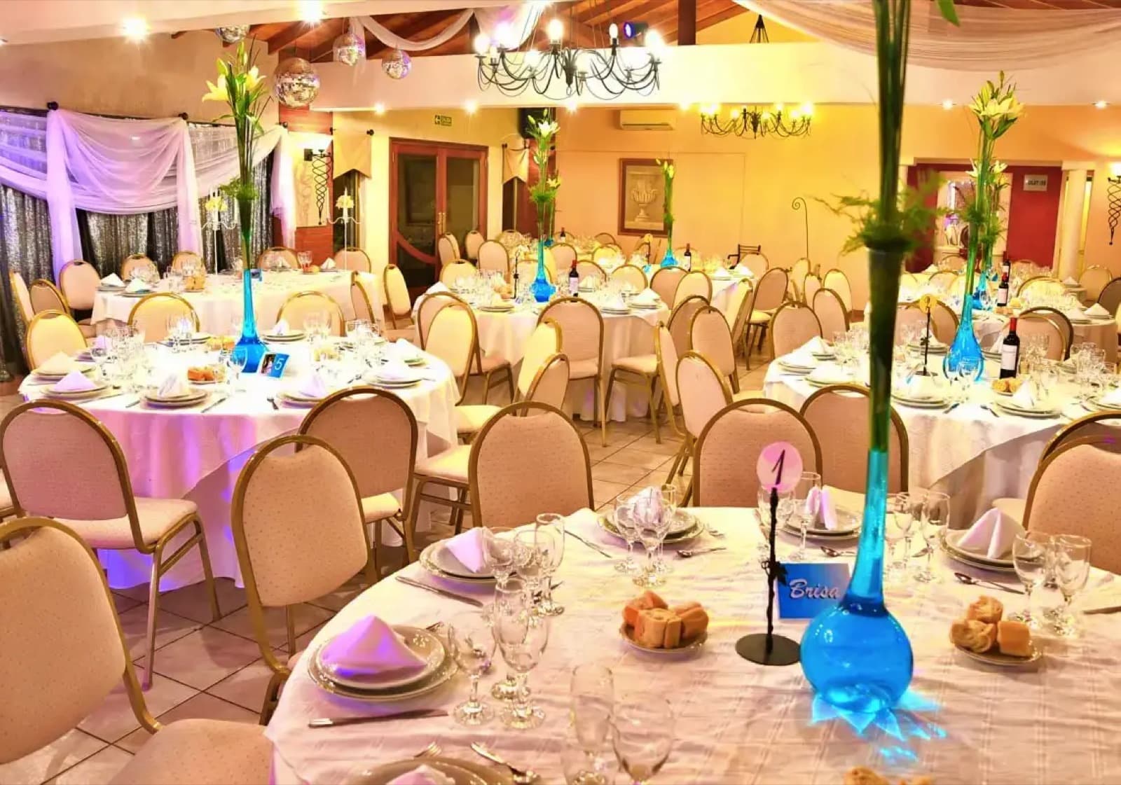 El salón puesto con mesas redondas, sillas tapizadas y centros de flores altos en floreros azules.