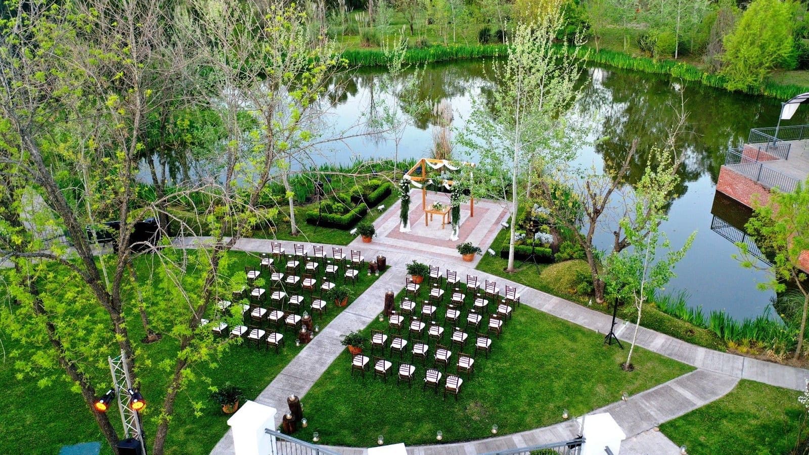 Vista aérea de la ceremonia montada junto a la laguna del predio, con las sillas vacías frente al arco de flores y el parque alrededor.