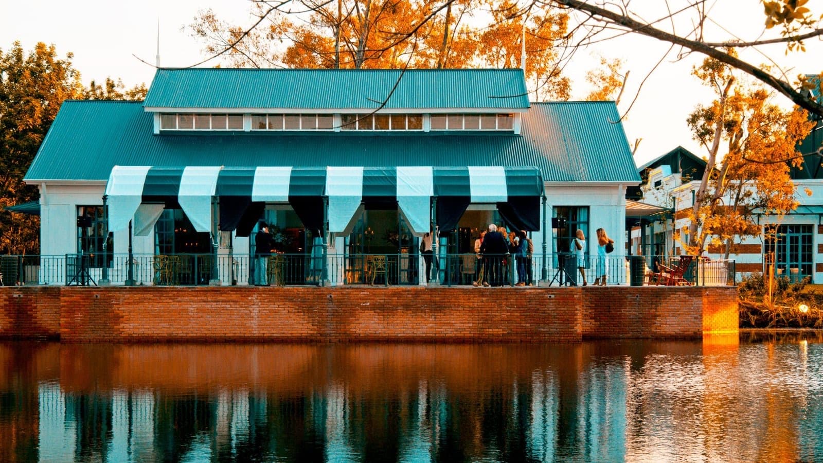 El Boat House de techo turquesa iluminado al atardecer, reflejado en la laguna, con los toldos a rayas abiertos.