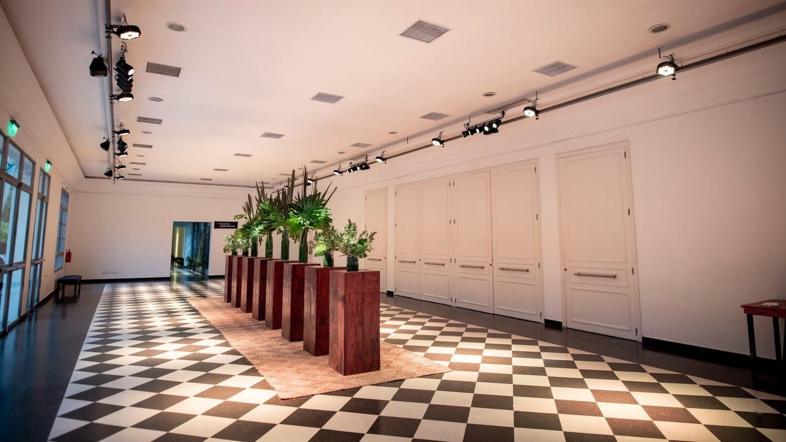 El salón vacío con el piso damero en blanco y negro, los canteros de plantas alineados al medio y las puertas blancas a un costado.