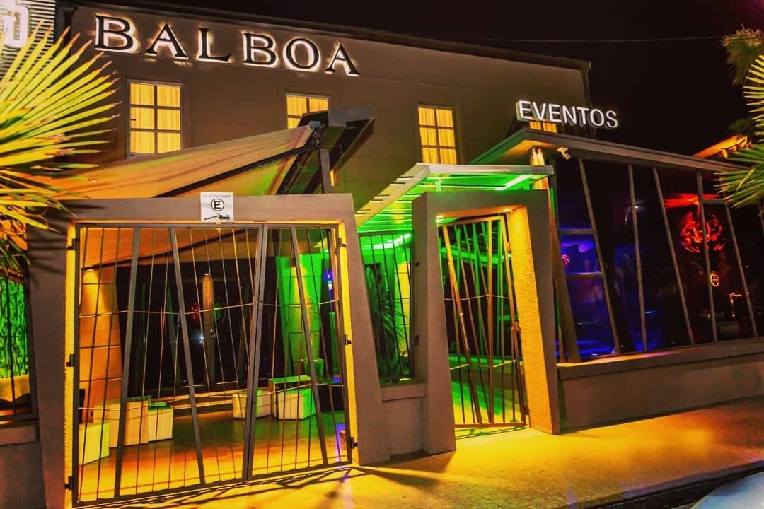 La fachada del salón de noche, con las letras Balboa Eventos sobre el frente, los portones abiertos y el interior iluminado de verde y amarillo.
