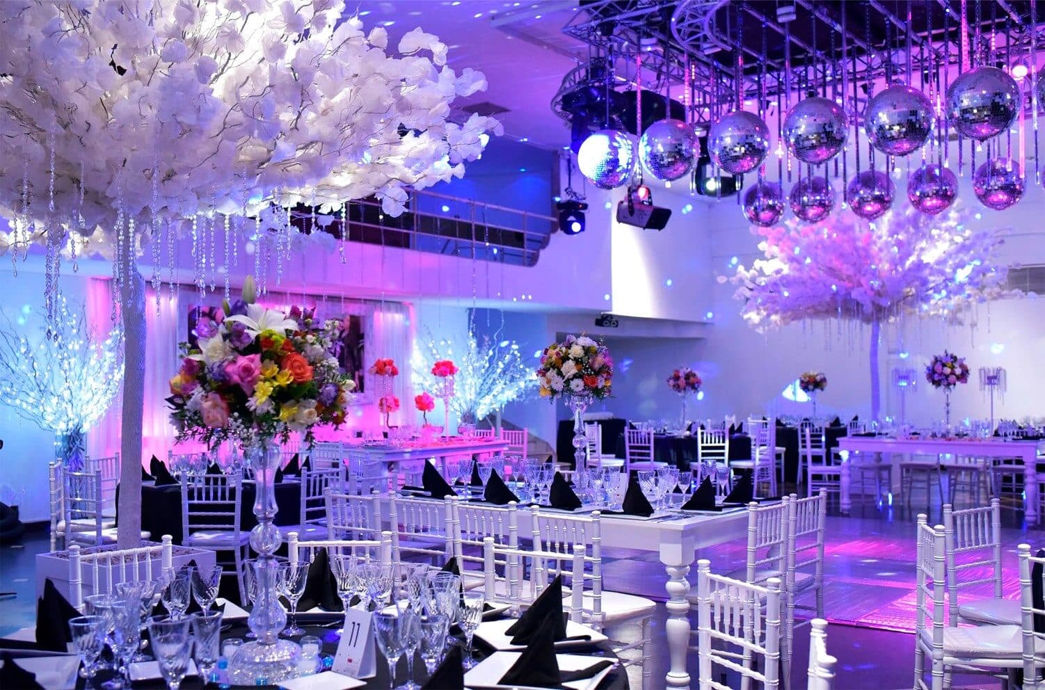 El salón ambientado en blanco y violeta, con árboles de flores blancas, centros de mesa altos con flores, sillas Chiavari blancas y bolas de espejo sobre la pista.