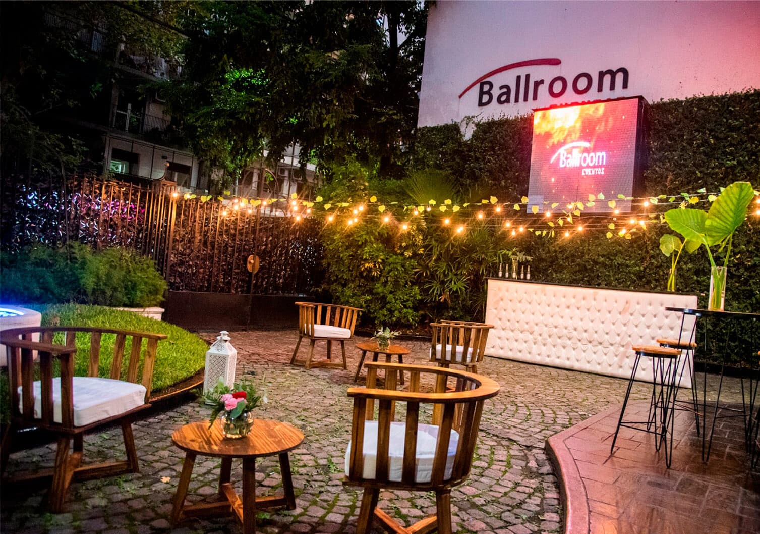 El patio al aire libre de noche, con sillones de madera alrededor de mesas bajas, guirnaldas de luces cálidas, canteros con verde y el cartel del salón al fondo.