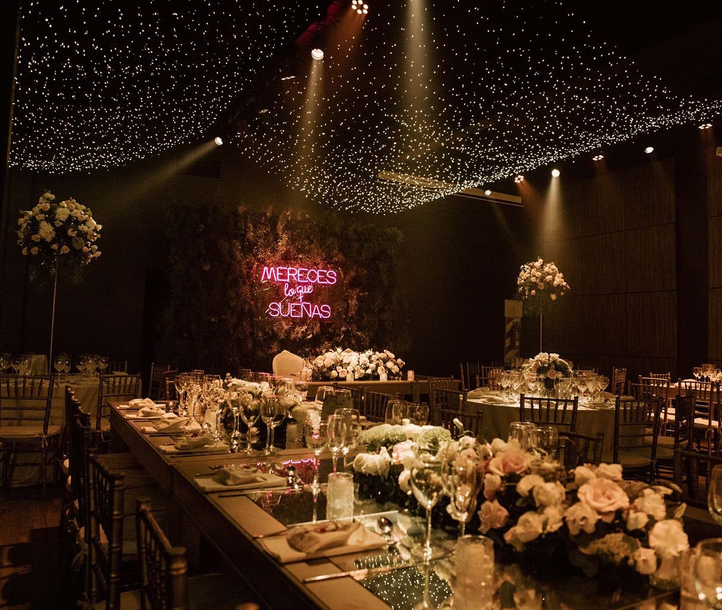 Una mesa larga puesta con flores blancas y velas, sillas de madera clara, un cielorraso de luces tipo estrellas y un cartel de neón en la pared del fondo.