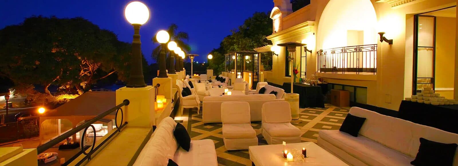 La terraza de noche, con sillones y pufs blancos alrededor de mesas bajas con velas, faroles de globo encendidos y la vista abierta al río detrás.