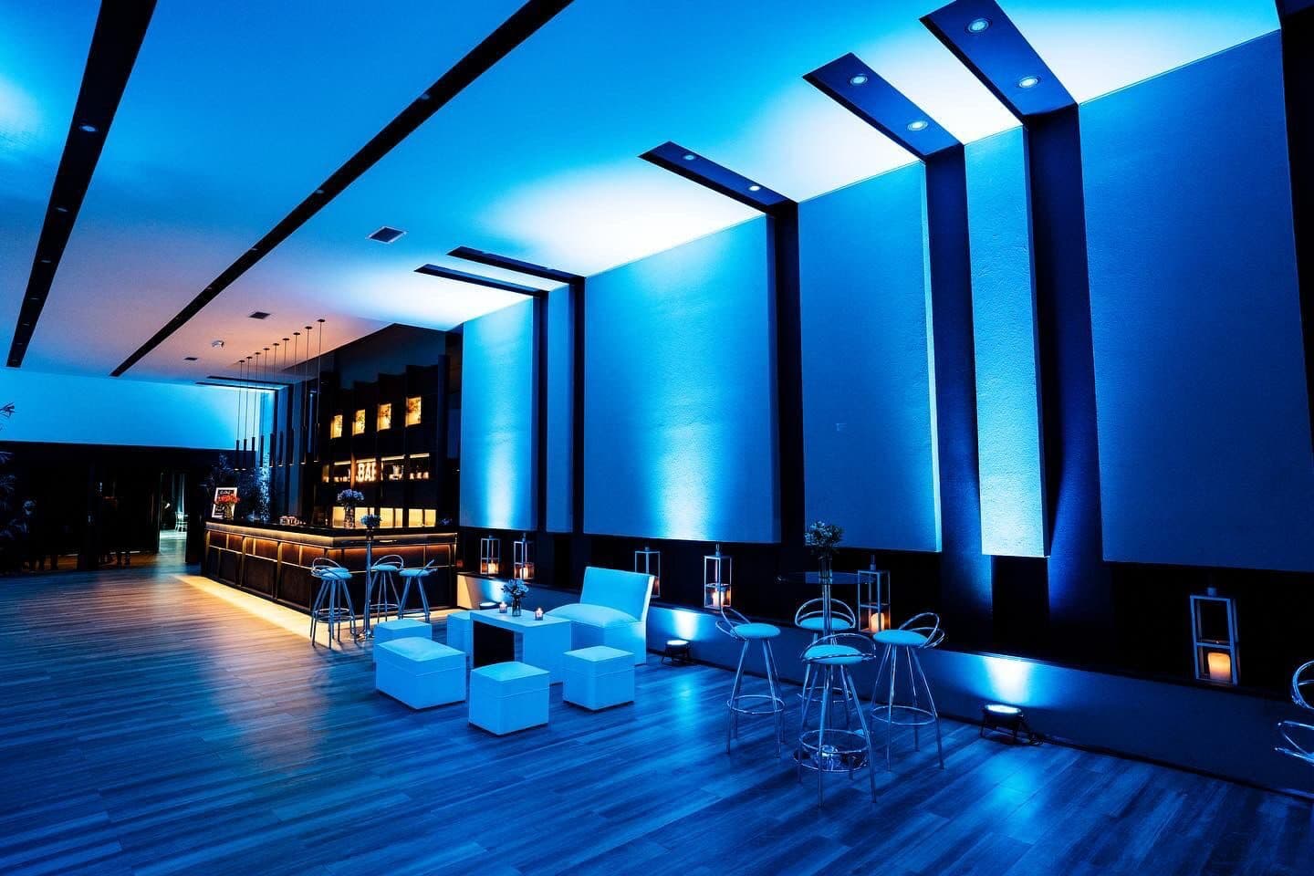 El salón vacío bañado en luz azul, con paneles retroiluminados en la pared, la barra al fondo y un living de puffs blancos con banquetas sobre el piso de madera.