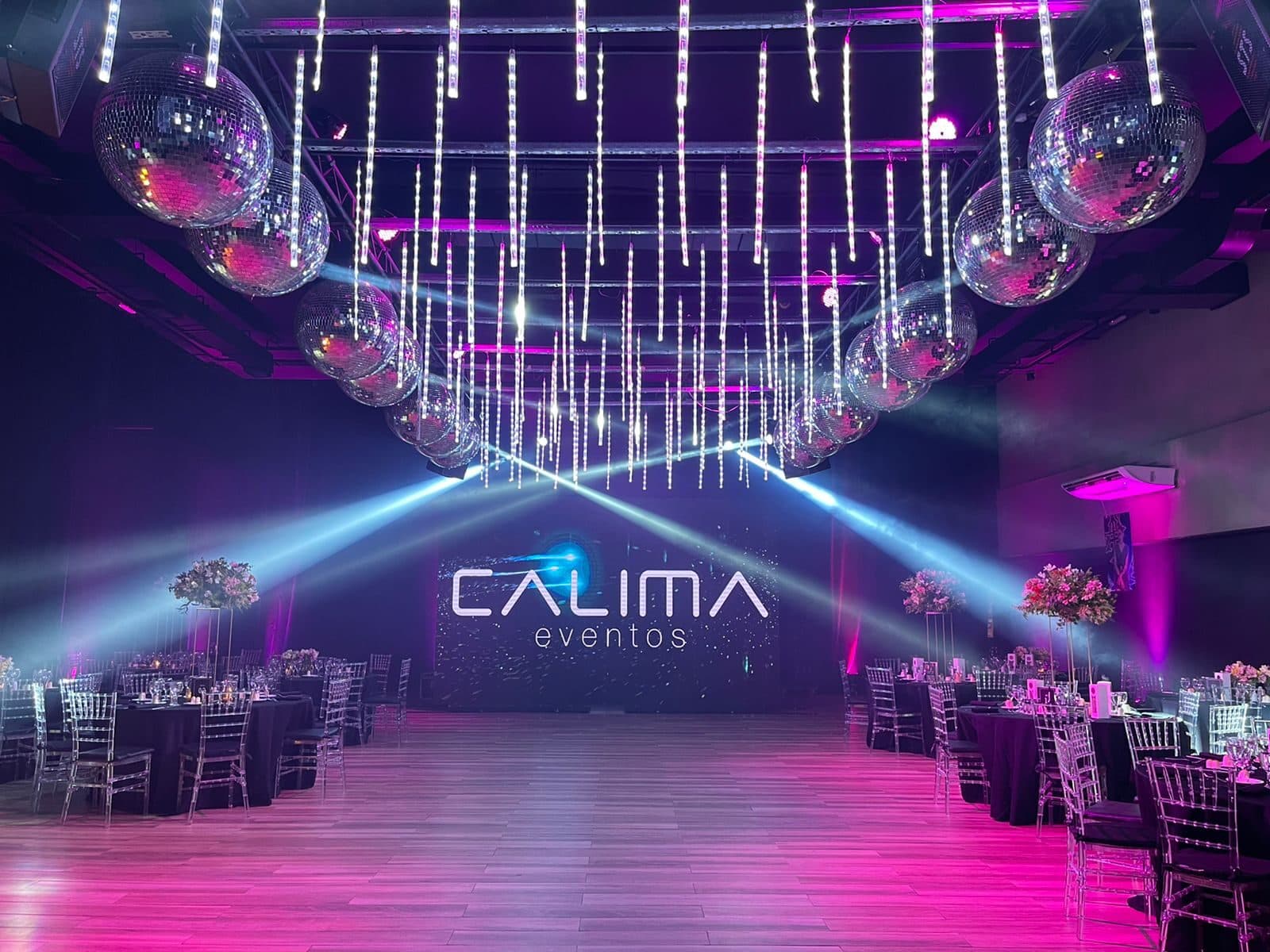 La pista de madera frente al cartel luminoso del salón, bajo bolas de espejo y tubos de luz colgantes, con las mesas de mantel negro y sillas transparentes alrededor.