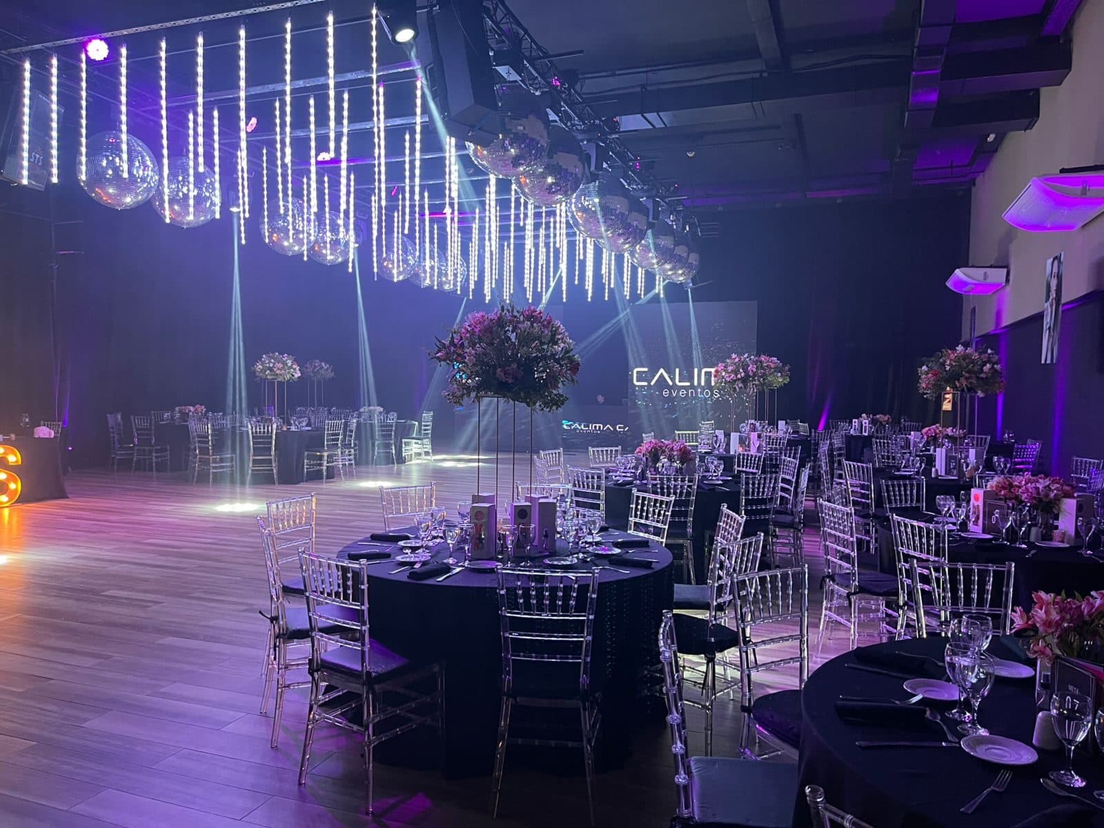 El salón armado para una fiesta con mesas redondas de mantel negro, sillas transparentes, centros de flores en pedestales altos y haces de luz cruzando la pista.