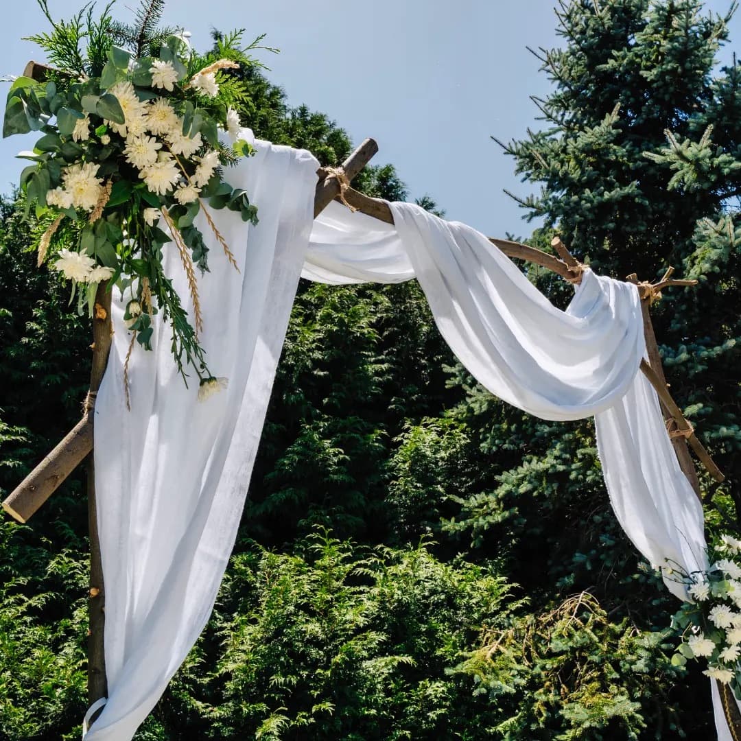 El arco de la ceremonia hecho con troncos, telas blancas y un ramo de flores en la esquina, contra los pinos del parque.
