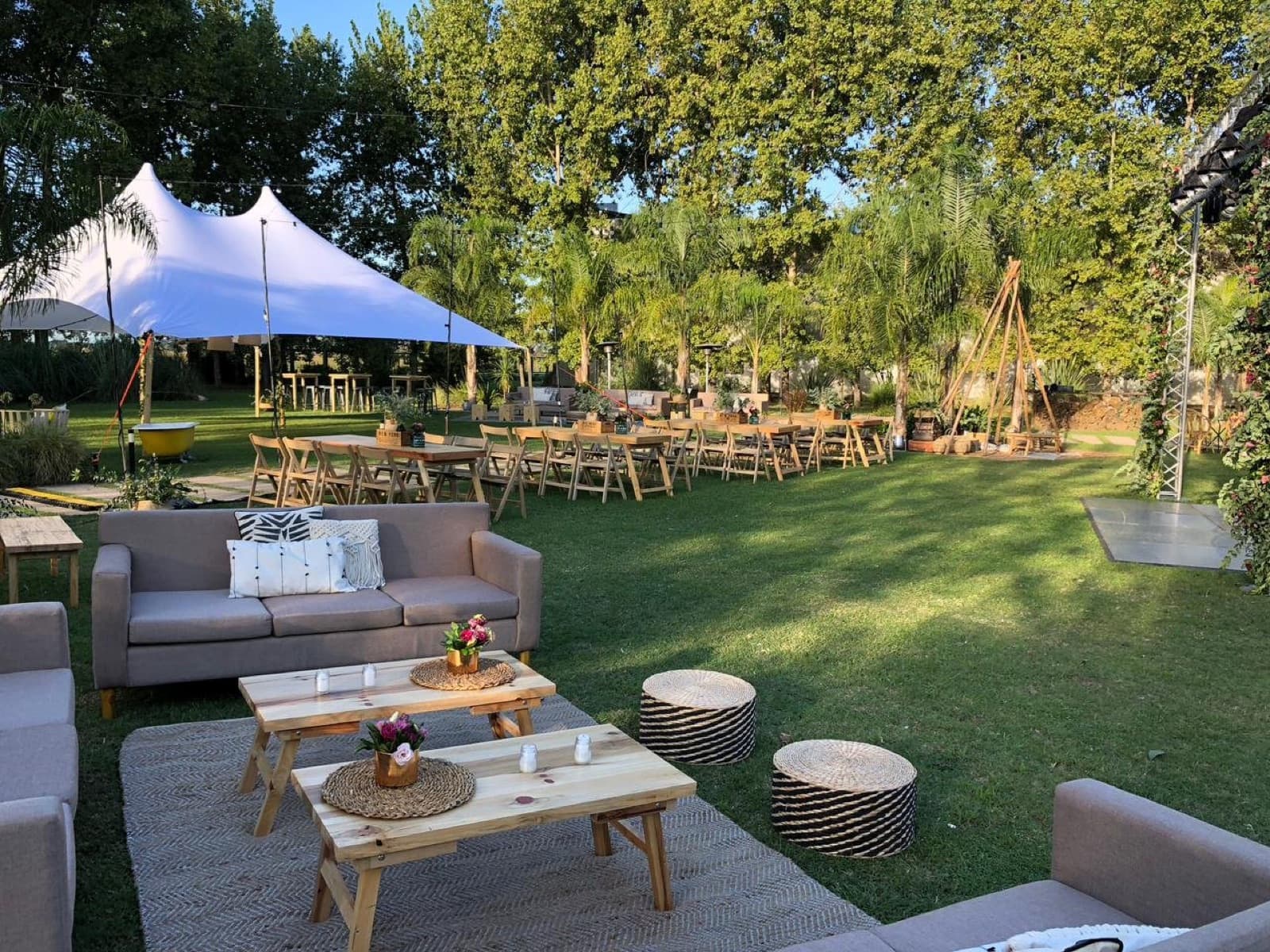 El parque con la carpa beduina blanca, las mesas largas de madera con sillas y los livings con alfombras sobre el césped, de tarde.