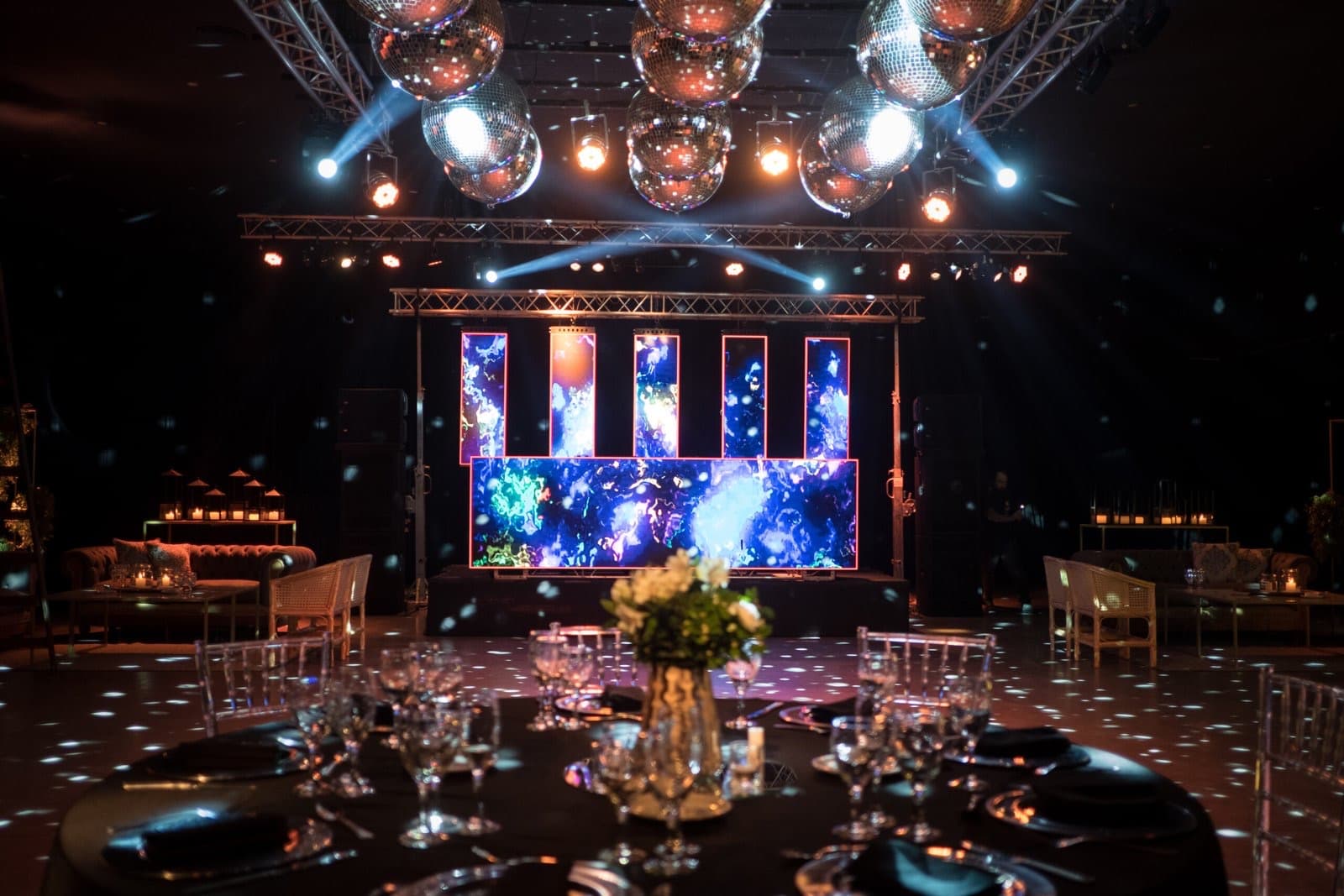 El salón de noche con bolas de espejos colgadas del techo, la pantalla LED sobre el escenario y una mesa redonda puesta en primer plano.
