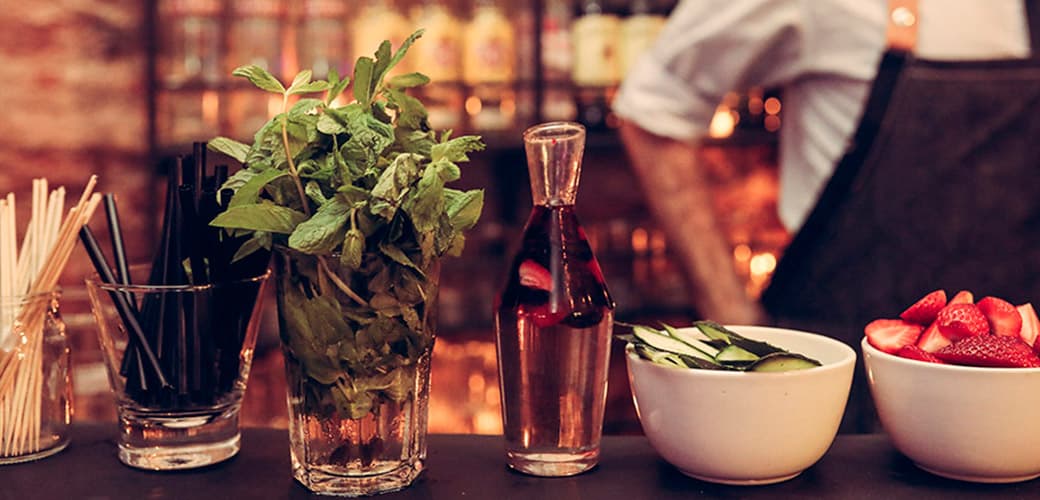 La barra de tragos con un vaso de menta fresca, una botella de sangría, bowls de pepino y frutillas y los sorbetes servidos, contra las botellas del fondo desenfocadas.