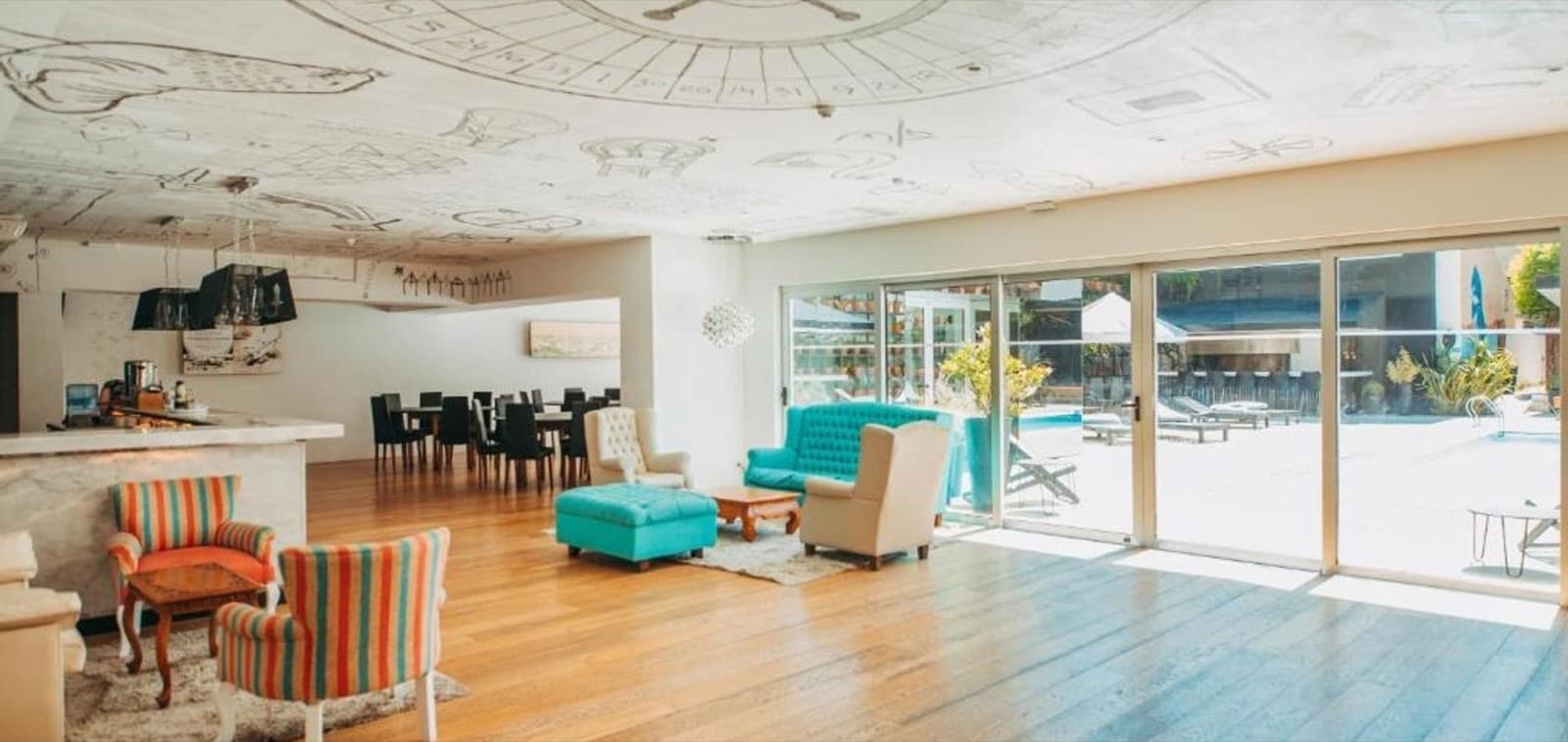 El living interior con sillones turquesa y a rayas sobre el piso de madera, el cielorraso dibujado y los ventanales a la pileta.