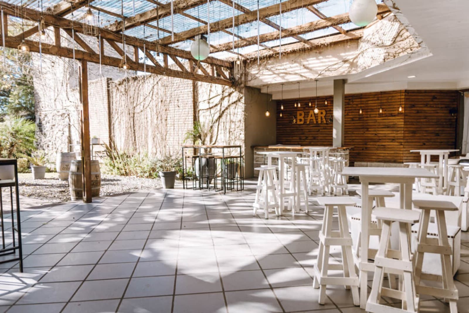 El patio techado con la pérgola de vidrio, mesas altas y banquetas blancas, y la barra de madera al fondo.