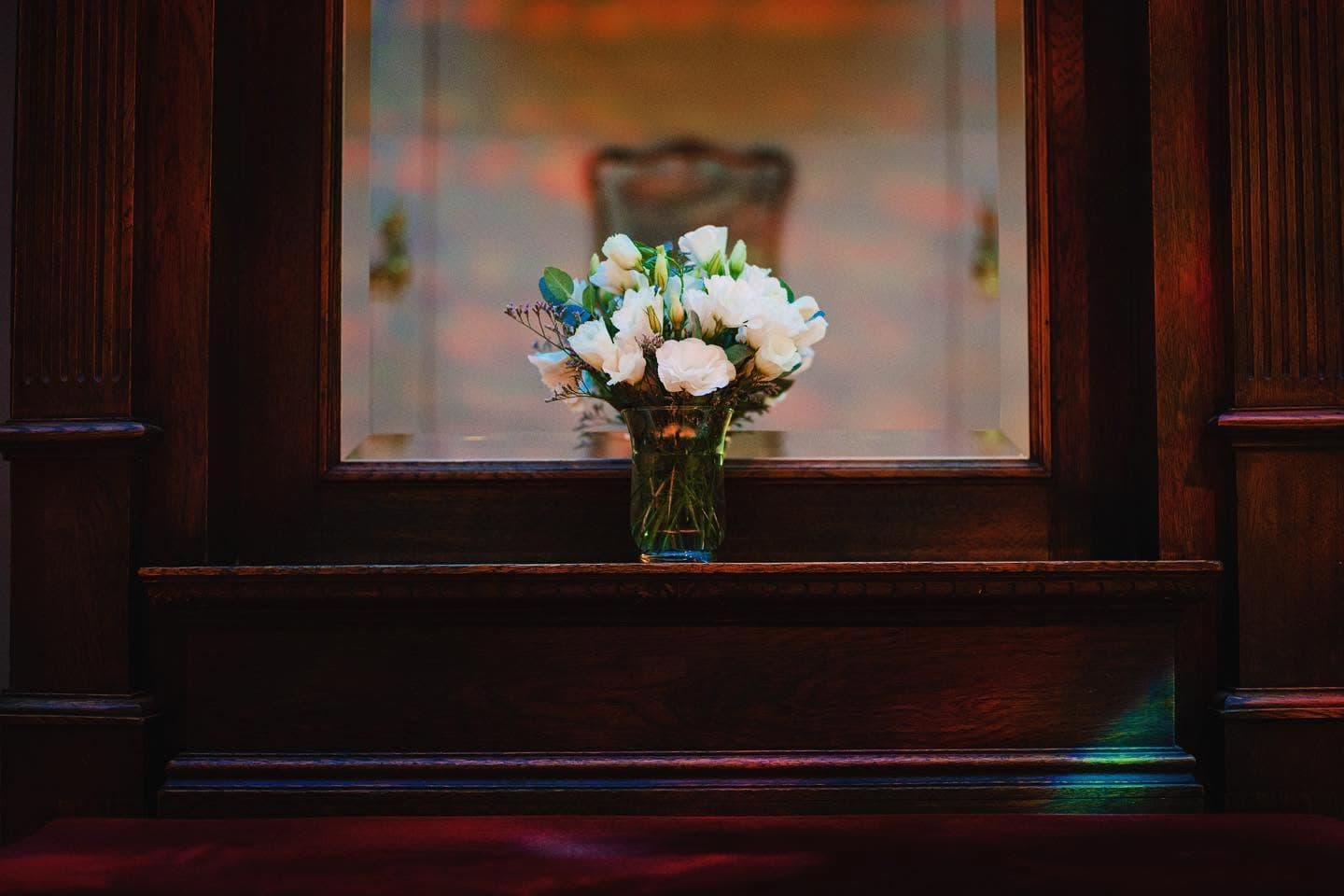 Un ramo de rosas blancas en un florero de vidrio, sobre el estante de un mueble de madera oscura con espejo, en penumbra.