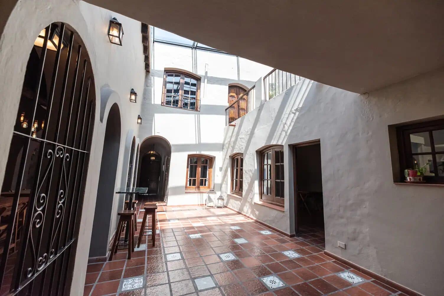 El patio de la casona con galería de arcos blancos, piso de baldosas rojas y calcáreas, faroles de hierro en las paredes y el techo vidriado dejando entrar el sol.