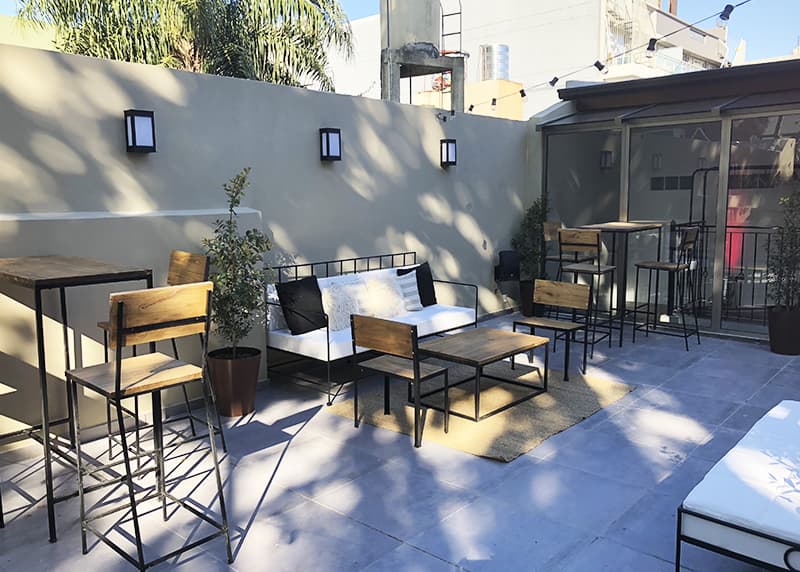 La terraza de día, con un sillón de hierro y almohadones, mesas altas de madera y macetas contra la pared clara, bajo guirnaldas de luces.
