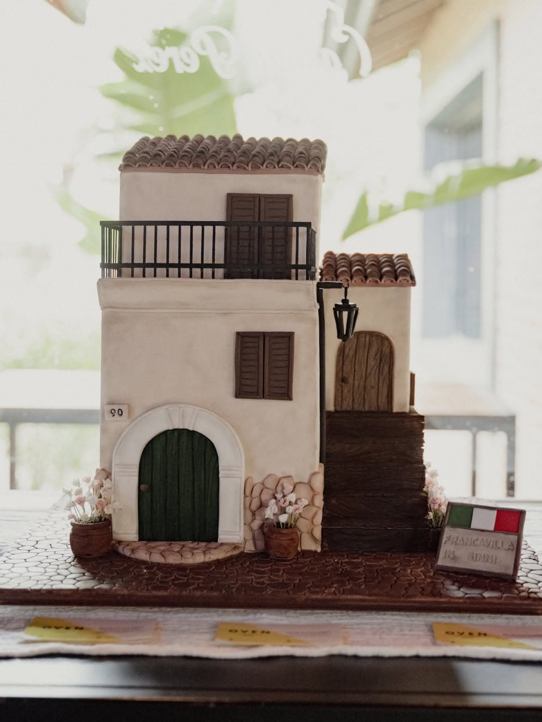 Una torta escultórica que reproduce una casa italiana de dos plantas, con tejas, balcón de hierro, puerta verde y macetas con flores, sobre un empedrado de azúcar.