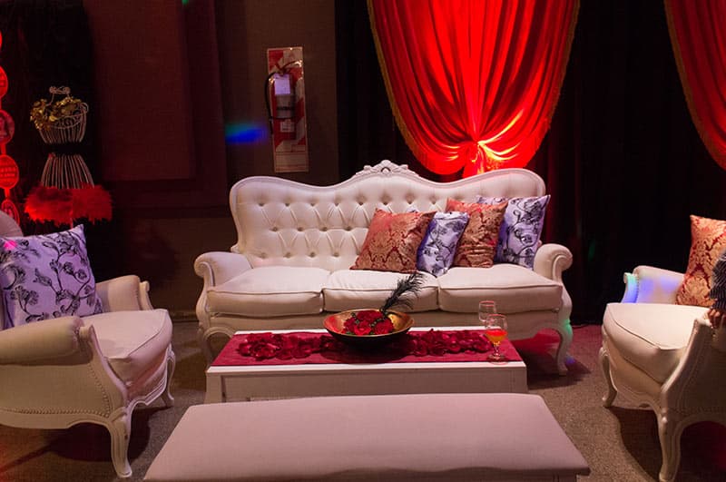 Un living armado en el salón: sofá y sillones blancos capitoneados alrededor de una mesa baja con pétalos rojos, contra un cortinado rojo iluminado.