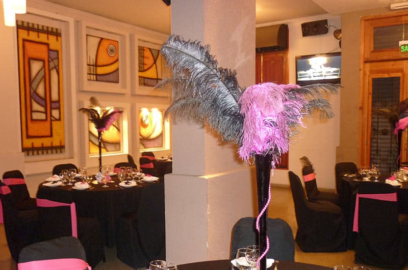 El salón puesto con mesas redondas de mantel negro, moños fucsia en las sillas y centros altos de plumas, con los cuadros de la pared iluminados.