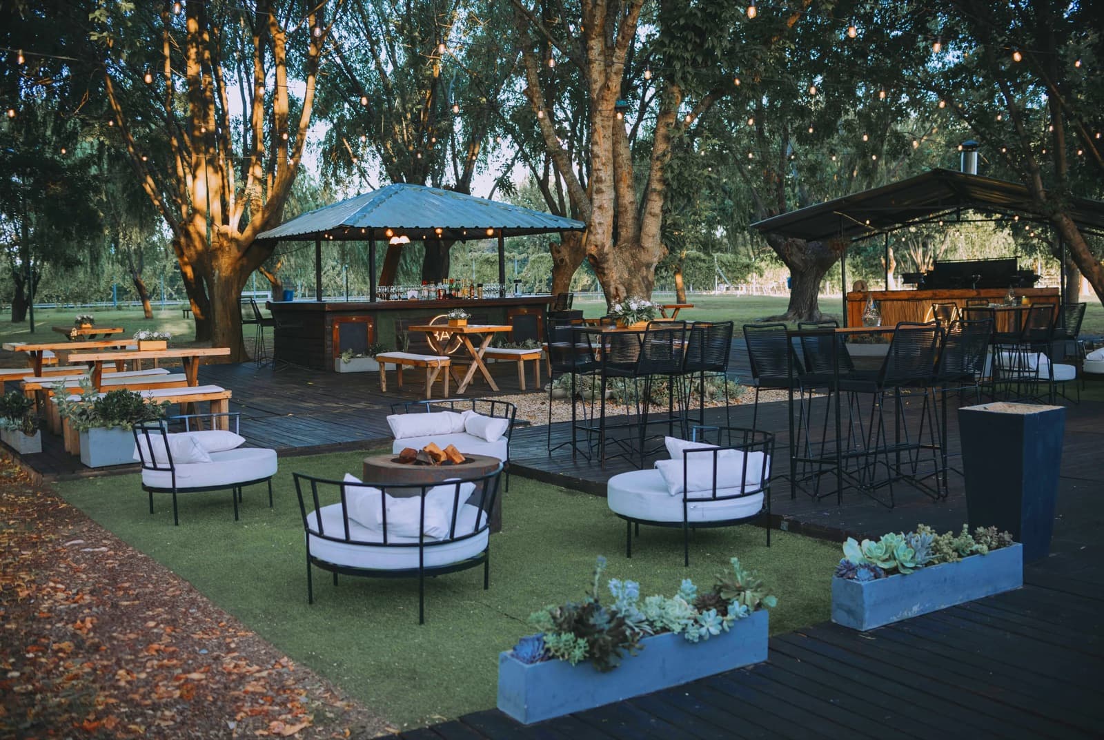 El deck exterior al atardecer con dos quinchos de chapa, uno de barra y otro de parrilla, mesas altas con sillas negras, sillones redondos alrededor de un fogón y guirnaldas de luces colgadas entre los árboles.