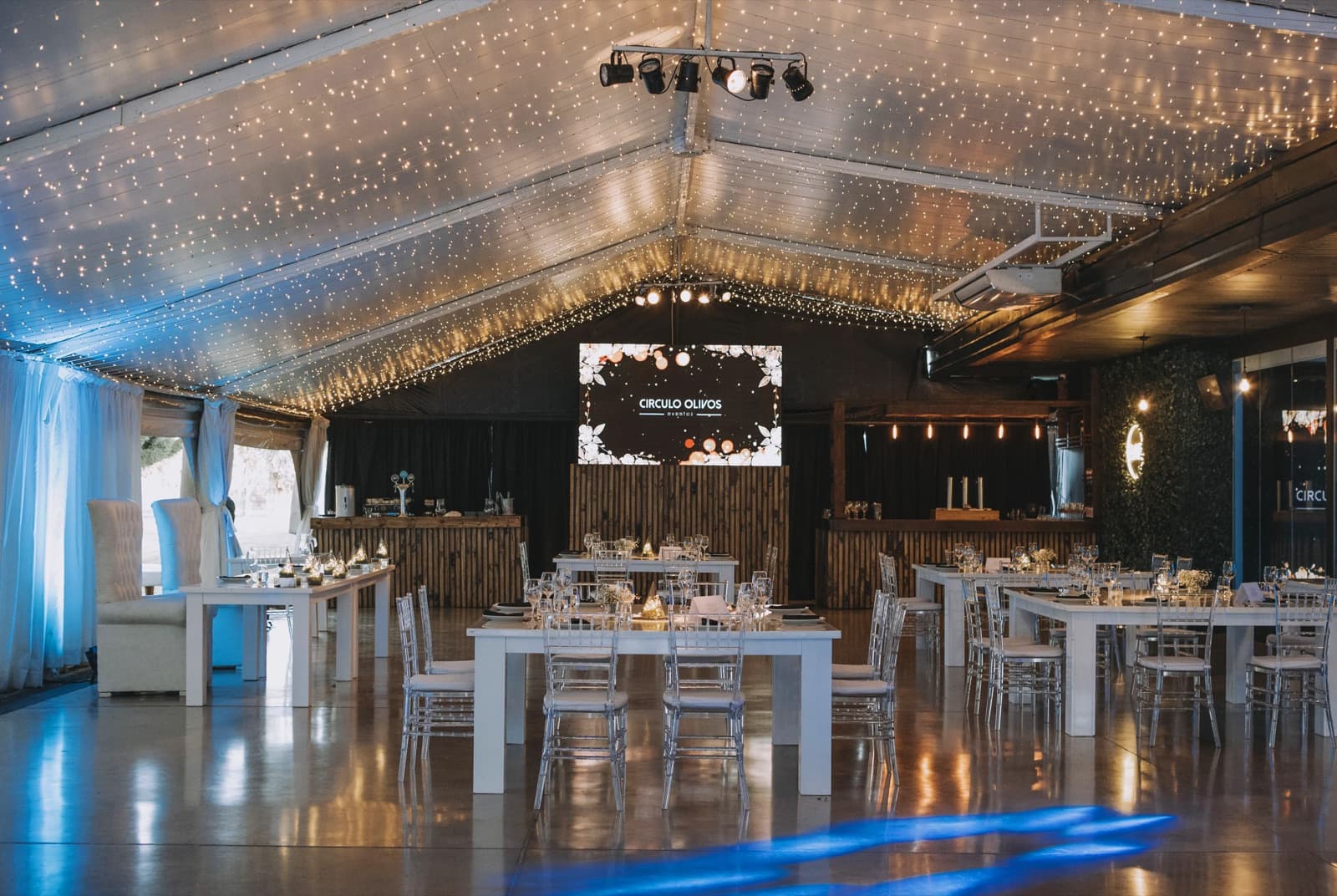 El salón semicubierto puesto y todavía vacío, con mesas blancas rectangulares y sillas Tiffany transparentes, el techo a dos aguas tapizado de luces cálidas y la pantalla con el logo de Círculo Olivos sobre la barra de madera del fondo.