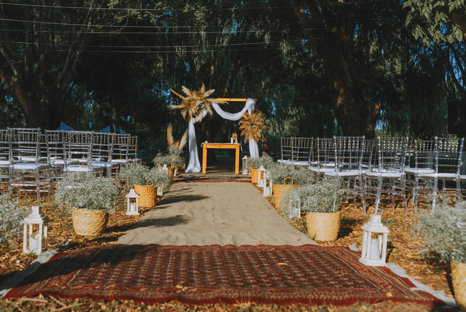 El sector de ceremonia armado bajo los sauces, con el camino de arpillera y una alfombra bordó entre canastos de paniculata, las hileras de sillas transparentes a los costados y un arco de madera con telas blancas y pampa seca al fondo.