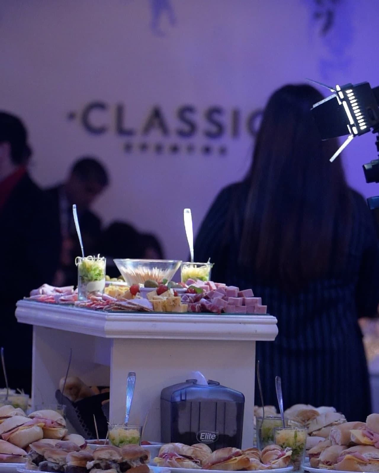 La mesa de fiambres y sándwiches servida en el salón, con la pared del nombre del lugar iluminada de violeta detrás.
