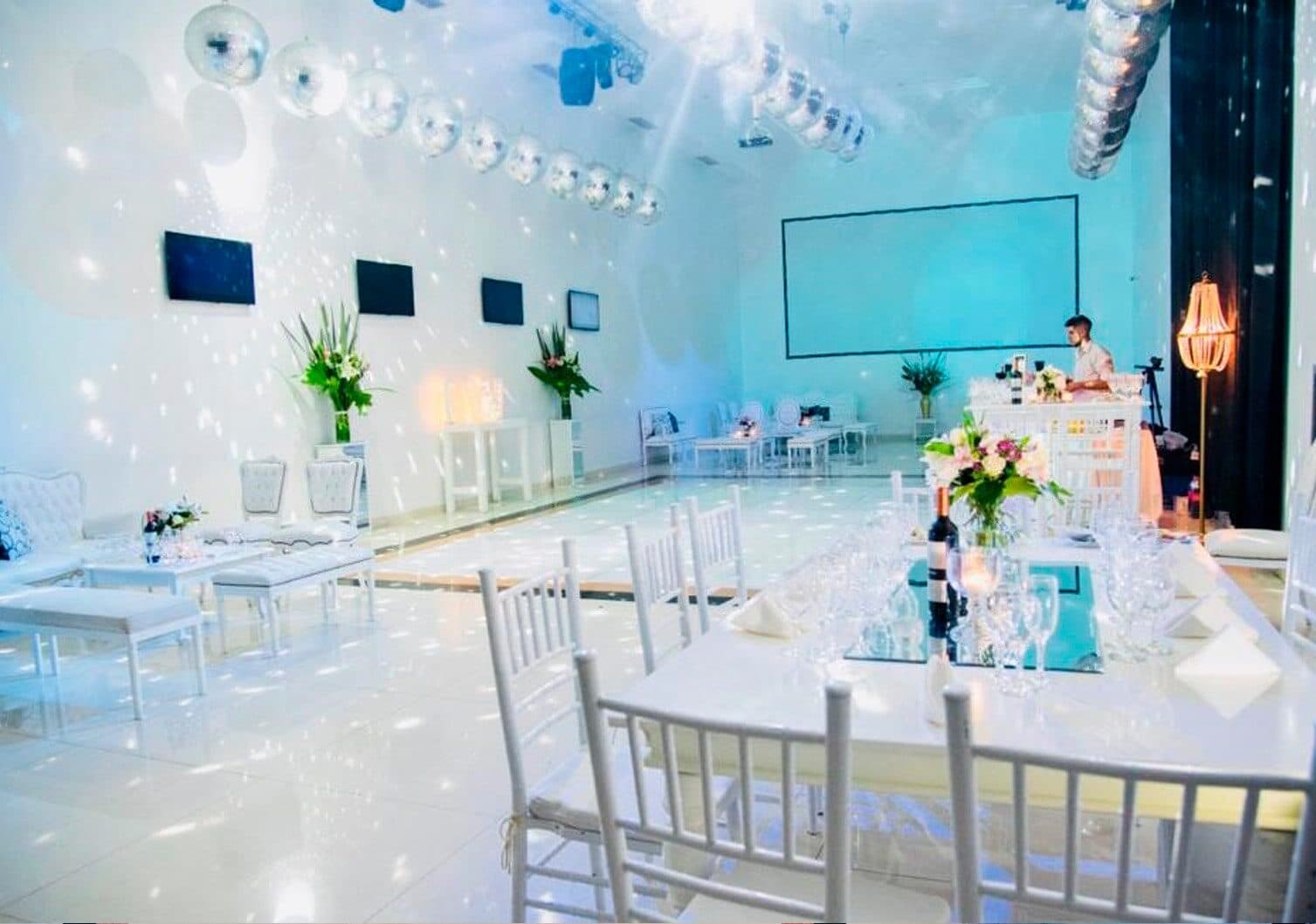 El salón armado con mesas de mantel blanco, sillas Chiavari y centros de flores, con guirnaldas de bolas de espejo cruzando el techo y una pantalla al fondo.