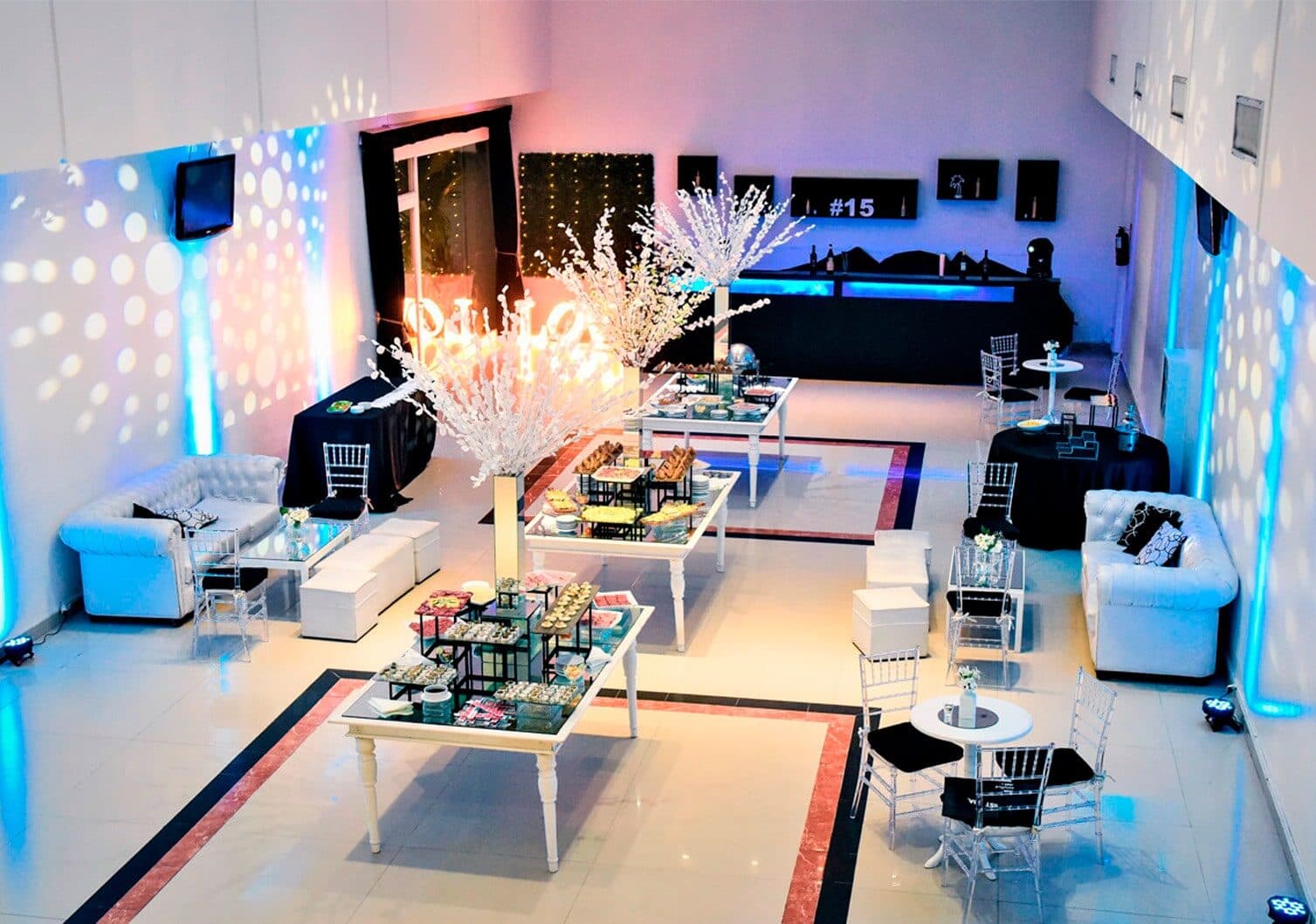 El salón en formato living, con sillones capitoné blancos, mesas bajas con bandejas, árboles de ramas blancas iluminadas y la barra negra al fondo.