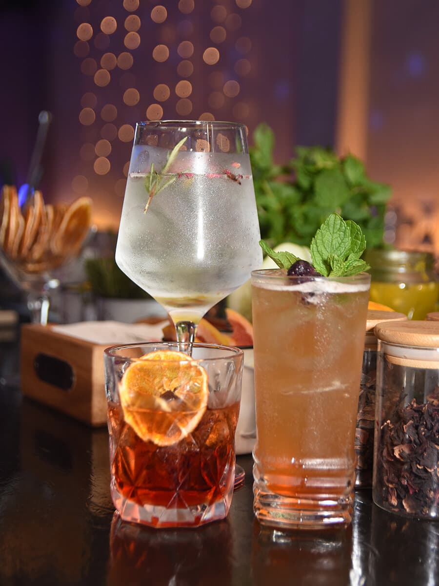 Tres tragos servidos sobre la barra del salón, con hielo, rodaja de naranja deshidratada y menta, contra las luces de la fiesta desenfocadas.