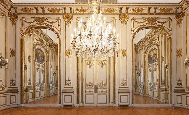 El salón de música vacío, con araña de caireles en el centro, molduras doradas sobre las paredes claras, espejos y puertas altas de doble hoja.