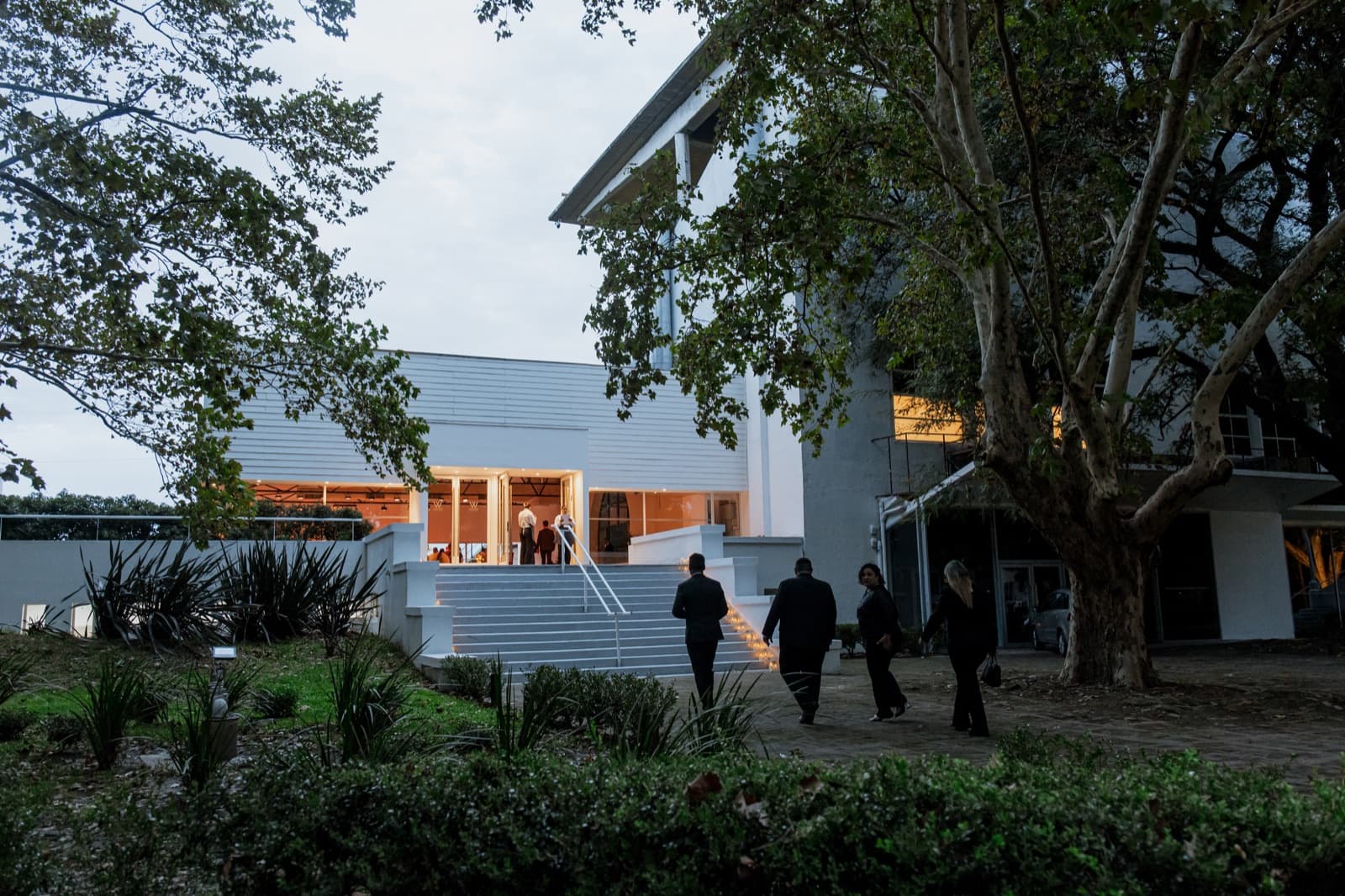 El edificio del salón al atardecer entre árboles grandes, con los ventanales encendidos de naranja, la escalinata de entrada con velas en los escalones y cuatro invitados de espaldas caminando hacia la puerta.