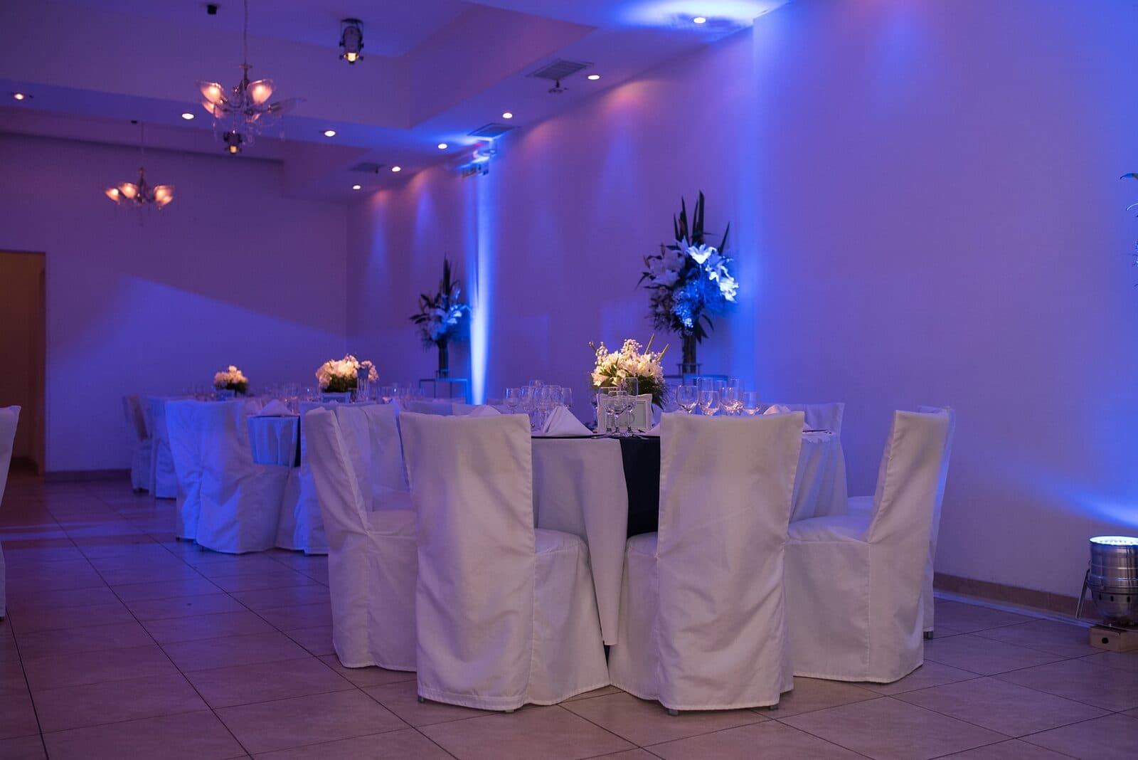 El salón armado con mesas redondas de mantel blanco, sillas vestidas, centros de flores altos y las paredes bañadas de luz azul.