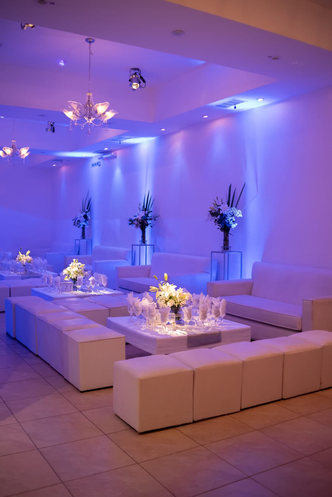 El salón en formato informal, con sillones y puffs blancos alrededor de mesas bajas con copas y flores, arañas de caireles y luz violeta sobre las paredes.