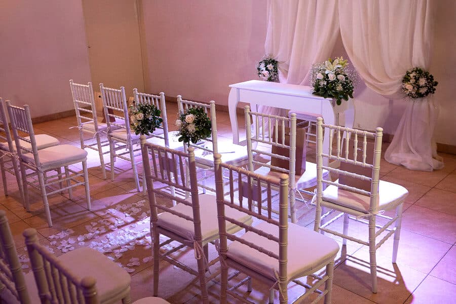 La ceremonia civil montada en el salón: hileras de sillas Chiavari blancas frente a una mesa vestida, con cortinado claro, ramos de flores y pétalos sobre el piso.