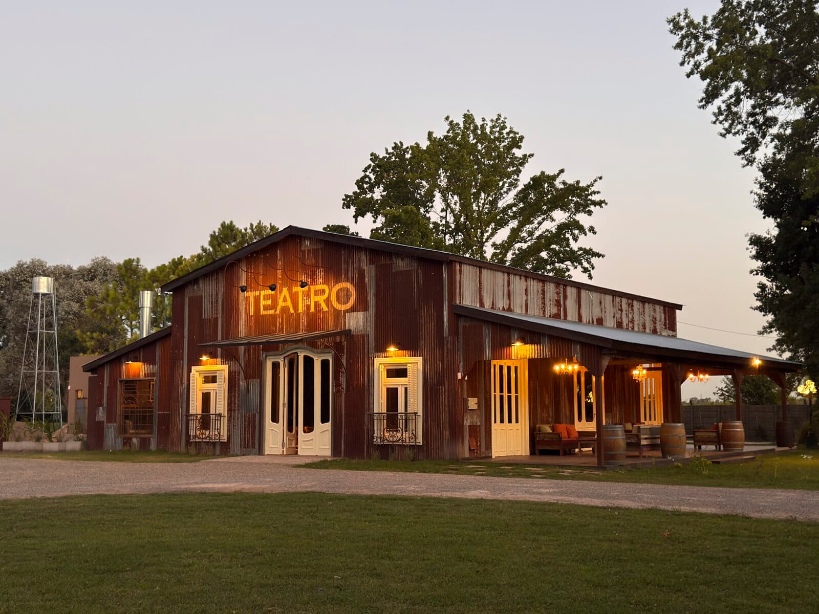 El teatro de madera al atardecer, con el cartel iluminado sobre la puerta, galería con faroles encendidos y árboles altos alrededor.