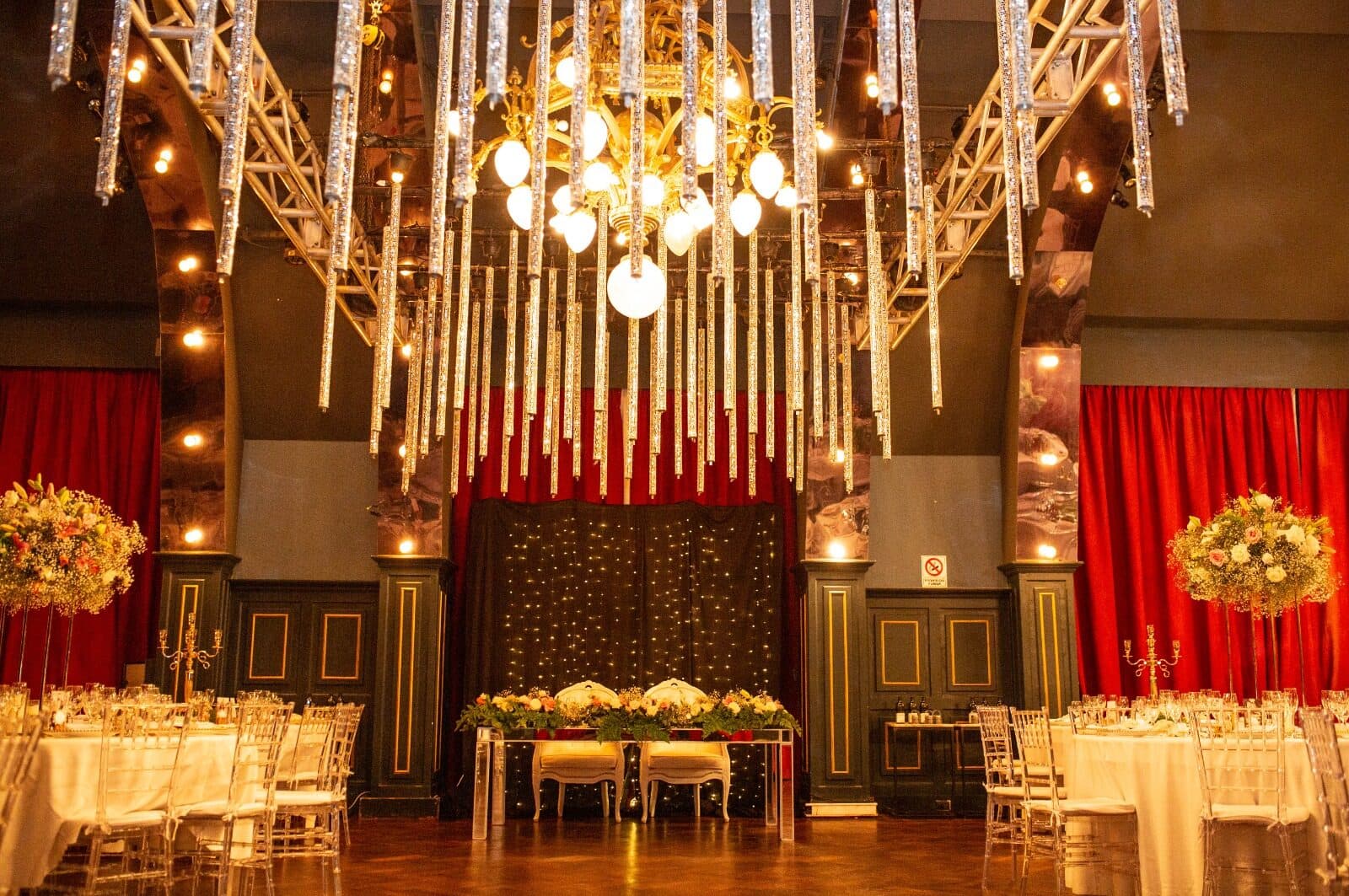 El salón principal armado para una fiesta, con una araña dorada y tubos de cristal colgando del techo, cortinas rojas, mesas de mantel blanco y centros de flores altos.